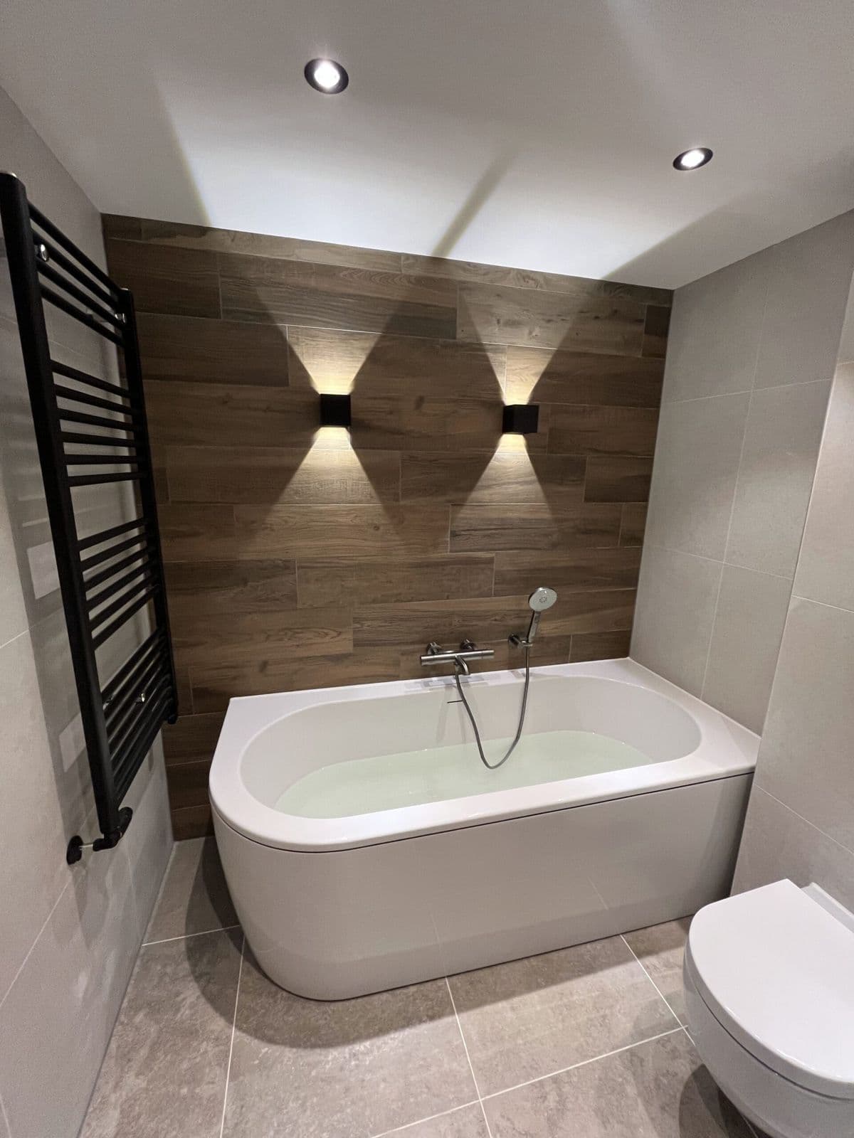 Badkamer met ligbad, wand in houtlook met wandlampen en hangtoilet